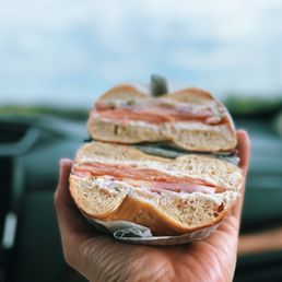 Lower East Side Bagel - A close-up of a bagel sandwich with smoked salmon, cream cheese, and possibly other ingredients, held in a person's hand inside a vehicle. The background shows a blurred sky and parts of the car's dashboard, emphasizing the focus on the bagel. The bagel appears freshly baked with a soft interior and a slightly toasted exterior.