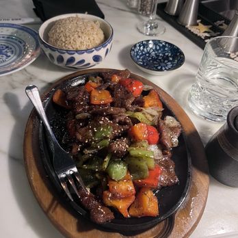 The 698 - Pepper steak - The image shows a sizzling beef stir-fry with mixed vegetables such as bell peppers, carrots, and onions on a cast-iron skillet placed on a wooden tray. Nearby, there is a bowl of rice, a glass of water, and small condiment containers. The setting appears to be a restaurant with a marble table surface, and various utensils and decorative items are visible in the background. - Sushi Bars Near Me - Columbus, Ohio