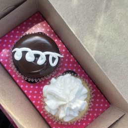 Two cupcakes are inside a cardboard box with pink polka dot paper. One cupcake has chocolate frosting with white icing decoration, the other has white frosting.