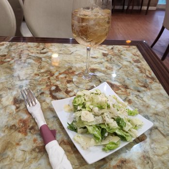 Hala Cafe - A glass of white wine with ice, a Caesar salad on a white plate, and a fork wrapped in a cloth napkin are on a marble table in a restaurant setting. - Mediterranean Near Me - Euless, Texas
