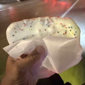 Voodoo Doughnut - Strawberry Go-Tart - A hand grips a donut decorated with white icing and small, multicolored sprinkles. The donut is partially wrapped in white paper. The background shows a parking lot with several cars and red taillights, indicating it is nighttime. The scene suggests the donut was purchased from a bakery or donut shop and is being enjoyed outdoors. - Donuts Near Me - Austin, Texas