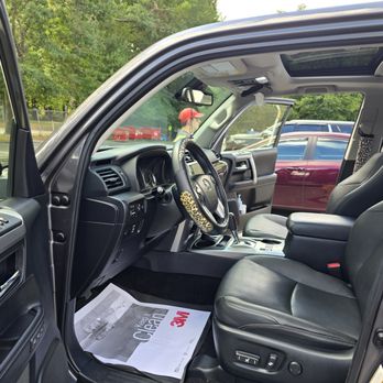 Innovative Auto Detailing - The photo captures the interior of a vehicle, featuring black leather seats, a dashboard with various controls, and a steering wheel wrapped in a leopard print cover. The vehicle has a sunroof, and a paper mat from 3M is on the floor. Outside the vehicle, there are parked cars and green trees, indicating an outdoor setting. The image is clear and well-lit, showing details of the vehicle's interior and surroundings. - Auto Detailing Near Me - Raynham, Massachusetts