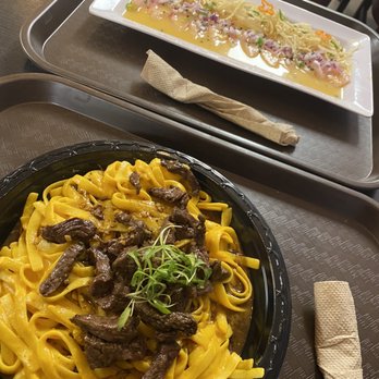 Mixturas Fusion - Steak fettuccini huancaina and ceviche sashimi - Two pasta dishes are shown on a restaurant tray: one with beef strips on yellow noodles, and another with seafood and noodle salad. - Peruvian Near Me - Orem, Utah