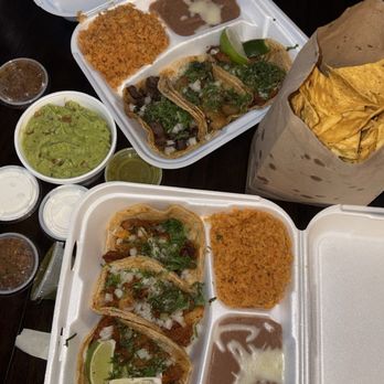 Edgewater Tacos - Taco dinners, chips and guac - Two styrofoam takeout containers hold tacos with lime wedges, rice, beans, and various sauces, accompanied by a paper bag of tortilla chips. - Tacos Near Me - Chicago, Illinois