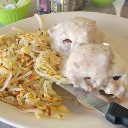 The image shows a white oval plate containing two Salisbury steaks covered with gravy and a portion of hash browns. The plate is placed on a table with a glass of water and condiments in the background, indicating a casual dining setting typical of a diner or American restaurant.