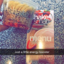 Shake smart acai bowl - A person holds a Shake Smart acai bowl container with sliced bananas and toppings, next to a Shake Smart menu on a granite counter.