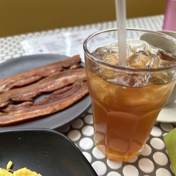 Sun Rey Cafe - The image shows a table setting with a tall glass of iced tea filled with ice cubes and a straw, a black plate with several strips of cooked bacon, and a white coffee cup with a saucer. The table has a white mesh surface, and there is a green napkin partially visible. The background is blurred, focusing on the food and drink items. - Breakfast & Brunch Near Me - Chicago, Illinois