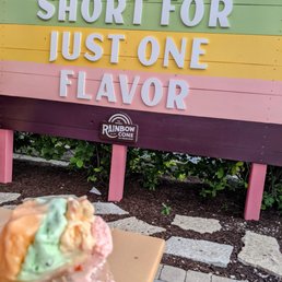 a person holding a donut - The image shows a colorful outdoor sign for Rainbow Cone ice cream shop, with horizontal stripes in orange, green, yellow, pink, and dark purple. The sign displays the quote, "Life's too short for just one flavor," in large, bold letters. In the foreground, a person holds a waffle cone with three scoops of ice cream in different colors, partially out of focus. The setting appears to be outside, with some greenery and stone paving visible at the bottom.