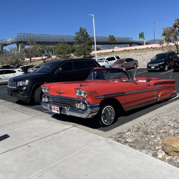 Twin Peaks - cool ride out front - A vintage red convertible car with chrome accents and a distinctive design is parked in a parking lot alongside contemporary SUVs and sedans. The car features a sleek, elongated body, prominent front grille, and wire-spoke wheels. The parking lot is situated near a hillside with trees, and a bridge structure is visible in the background under a clear blue sky. The scene captures a contrast between classic and modern vehicles in an outdoor setting. - American Near Me - Albuquerque, New Mexico