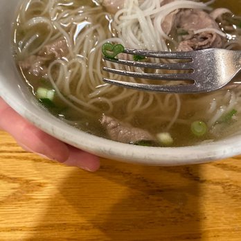 Pho Duy - Rare steak and Brisket - A bowl of Vietnamese pho contains clear broth, rice noodles, slices of beef, and chopped green onions. A fork is positioned above the bowl, holding a small piece of green onion shaped like a heart. The bowl is held by a hand over a wooden table, with the focus on the food and utensil. The image emphasizes the ingredients and presentation of traditional Vietnamese pho. - Vietnamese Near Me - Greeley, Colorado
