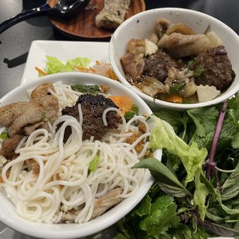 84 Viet - Hanoi Style Grilled Pork Vermicelli with dipping broth. Pork and sauce added to the vermicelli - The image shows two bowls of Vietnamese noodle soup with meat, vegetables, and herbs on a dark table. One bowl contains rice noodles, grilled meat, and vegetables, while the other has chunks of meat and vegetables in broth. Fresh herbs and leafy greens are arranged beside the bowls, along with a small side dish and a glass of water. The setting appears to be in a restaurant with a modern interior. - Vietnamese Near Me - Berkeley, California