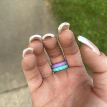 Oasis Nails & Spa - Messy and she got the polish everywhere - A close-up of a hand with manicured nails and two pastel-colored rings, held outdoors near a grassy area and a sidewalk. - Nail Salons Near Me - Bloomington, Illinois