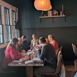 Several individuals sit around a wooden table in a restaurant with dark walls and large windows, illuminated by hanging paper lanterns.