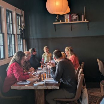 Catbird - Several individuals sit around a wooden table in a restaurant with dark walls and large windows, illuminated by hanging paper lanterns. - Cocktail Bars Near Me - South Berwick, Maine