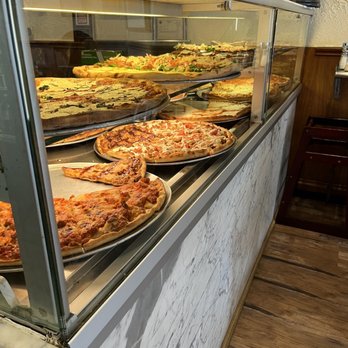 La Strada Restaurant - Pizza pizza! - A glass display case contains several pizzas with various toppings, including vegetables and meats, arranged on metal trays. The case is part of a restaurant interior with a marble-like counter and wooden flooring, and some framed certificates or menus are visible in the background. - Pizza Near Me - Millburn, New Jersey