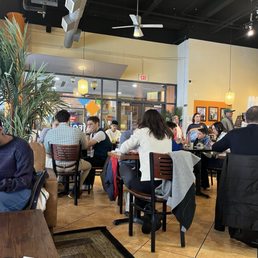 A crowded restaurant interior with diverse patrons seated at tables, some engaged in conversation, and a glass door entrance visible in the background.