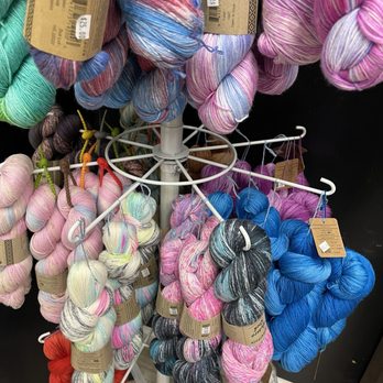 Needful Things - Multiple skeins of yarn in various colors and patterns are arranged on a white spinning rack in a craft or yarn store. - Knitting Supplies Near Me - Turlock, California