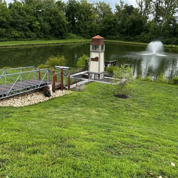 Piazza Messina - Lighthouse - A pond with a fountain is surrounded by lush green grass and trees. A small lighthouse structure is situated near the water, with a boat on a dock nearby. The scene includes a metal bridge leading to the lighthouse and a white picket fence around the structure, with the background filled with dense trees. - Venues & Event Spaces Near Me - Cottleville, Missouri