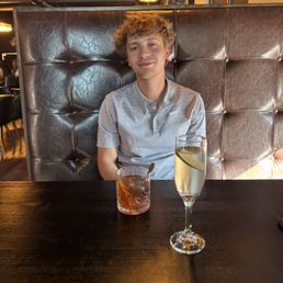A young man with curly, light brown hair and fair skin is seated at a dark wooden table in a restaurant. He wears a light gray, striped polo shirt and has a neutral expression. In front of him are two drinks: a short glass with a dark-colored beverage and a straw, and a tall champagne flute with a clear liquid and a lemon slice. The background features a padded leather booth, warm lighting fixtures, and a modern interior design.