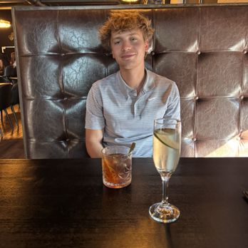 Citywalk Eatery - A young man with curly, light brown hair and fair skin is seated at a dark wooden table in a restaurant. He wears a light gray, striped polo shirt and has a neutral expression. In front of him are two drinks: a short glass with a dark-colored beverage and a straw, and a tall champagne flute with a clear liquid and a lemon slice. The background features a padded leather booth, warm lighting fixtures, and a modern interior design. - American Near Me - Cedar Rapids, Iowa