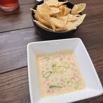 The Jack - A white square bowl filled with a creamy, pale-colored dip topped with sliced green onions is positioned on a wooden table. Behind the bowl, a black dish contains tortilla chips. Two drinks, one red and one clear, are partially visible in the background. The setting appears to be a casual dining environment. - American Near Me - Camden, Maine
