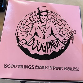 Voodoo Doughnut - The image shows a pink box with a black illustration of a person wearing a suit, hat, and sunglasses, with arms extended. The person is surrounded by multiple boxes arranged in a circular pattern. Below the illustration, the text reads "Good things come in pink boxes." The design is stylized and appears to be a logo or branding element, with a focus on the pink color and the message conveyed by the text. - Donuts Near Me - Denver, Colorado