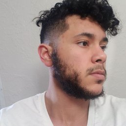 Got a medium fade from Thomas earlier today. I love it! - A young man with dark, curly hair and a full beard is captured in a side profile shot. He has light skin and is wearing a white V-neck shirt. The background features a plain, textured wall. The lighting is soft and even, highlighting his facial features and hair texture.