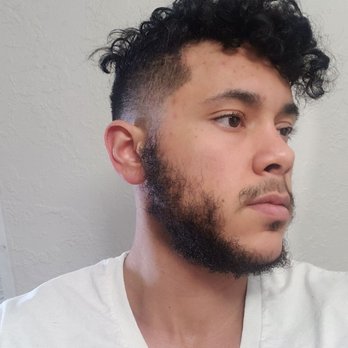Sir Men’s Salon - Got a medium fade from Thomas earlier today. I love it! - A young man with dark, curly hair and a full beard is captured in a side profile shot. He has light skin and is wearing a white V-neck shirt. The background features a plain, textured wall. The lighting is soft and even, highlighting his facial features and hair texture. - Men's Hair Salons Near Me - Albuquerque, New Mexico