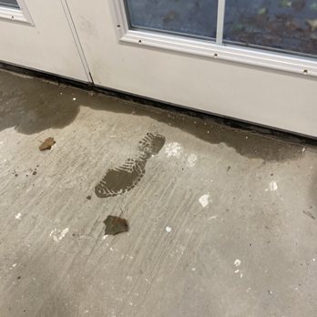 Solid Rock Builders - water coming into our mudroom because he didn't using a sill pan, or silicone to set the door, which is so not plum or level. HACK!!! - A wet footprint is seen on a concrete floor near a glass door, with some leaves and dirt scattered around. - Masonry/Concrete Near Me