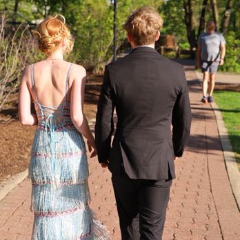 Peaches Boutique - Jovani 38846 - A woman in a colorful, fringed dress and a man in a black suit walk together on a park pathway, with a person in casual clothing in the background. - Women's Clothing Near Me - Chicago, Illinois