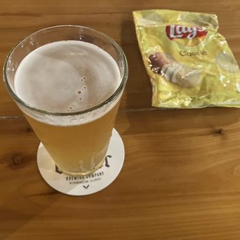 Casper Brewing Company - Citrus Wheat - A tall glass of beer with foam on top sits on a coaster on a wooden table, with a bag of Lay's Classic potato chips beside it. - Breweries Near Me - Bloomington, Illinois