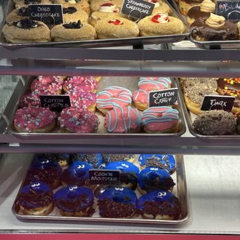 Hurts Donut - Yeast donut case - I was amused by the Cookie Monster donut. - A glass display case contains various decorated donuts with labels such as Oreo cheesecake, strawberry cheesecake, cotton candy, and cookie monster. The donuts are colorful and themed for different flavors. - Donuts Near Me - Cincinnati, Ohio