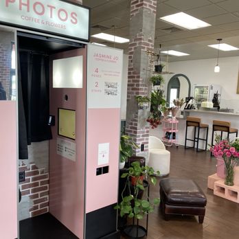 Jasmine Jo - The image shows a photo booth with a pink exterior and black curtain, located inside Jasmine Jo Coffee & Flowers. The booth has a screen and a camera, with a sign indicating it offers four poses and two strips. The surrounding area includes potted plants, a brown leather ottoman, and floral decorations, with a cozy interior featuring brick accents, indoor plants, and a counter in the background. - Coffee & Tea Near Me - Tempe, Arizona