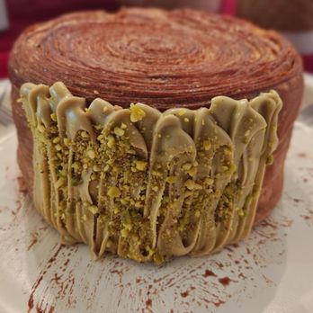 Baker 305 - A pastry cake features a spiral pattern on the top and is decorated with pistachio-flavored frosting on the side, topped with chopped pistachios. The cake is placed on a white surface with some chocolate or cocoa powder smudges around it. - Bakeries Near Me - Miami, Florida