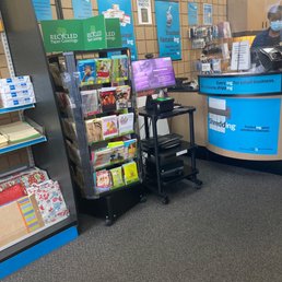 The image shows the interior of a postal service store, featuring a greeting card display rack with various cards, a checkout counter with a staff member wearing a face mask, and multiple signs advertising services such as recycling paper greetings, LED paper greetings, and notary services. The background includes a wall with informational posters and a computer monitor on a small cart. The floor is carpeted, and the overall setting is typical of a retail postal and shipping location.