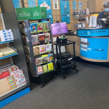 The UPS Store - The image shows the interior of a postal service store, featuring a greeting card display rack with various cards, a checkout counter with a staff member wearing a face mask, and multiple signs advertising services such as recycling paper greetings, LED paper greetings, and notary services. The background includes a wall with informational posters and a computer monitor on a small cart. The floor is carpeted, and the overall setting is typical of a retail postal and shipping location. - Shipping Centers Near Me - Hollis, New York