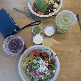 CAVA - Lemon chicken bowl & Harissa Avocado Bowl - Two bowls of salad with various vegetables, cheese, and toppings, along with two drinks and small containers of sauces or dressings on a wooden table. - Mediterranean Near Me - Cedar Park, Texas