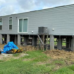 A long shot of a mobile home elevated on concrete blocks and metal supports, with construction debris and equipment on the gr...