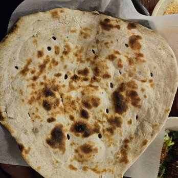 Sheeba Restaurant - Rashush - A sizable, round naan bread with uneven browning and charred spots is placed on a paper-lined tray. The bread appears freshly baked, with a slightly irregular shape and small holes. Part of a plate with food is visible in the background, indicating a dining setting. - Middle Eastern Near Me - Dearborn, Michigan