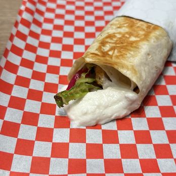 Alpha's Shawarma - A wrap sandwich with a toasted, golden-brown tortilla, filled with lettuce, grilled chicken, and sauce, partially wrapped in white paper, placed on a red and white checkered paper liner on a tray. The setting appears to be a casual dining environment. - Lebanese Near Me - Toronto, Ontario