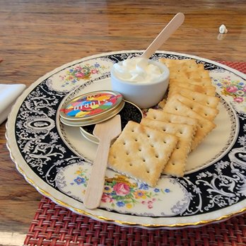 Frenchish - Caviar, creme fraiche, saltines. - A decorative porcelain plate with floral and black patterns contains saltine crackers, a small bowl of cream cheese with a spoon, and a wooden spreader. The plate is placed on a wooden table with a red woven placemat nearby, and a white napkin is partially visible on the left side of the image. - French Near Me - Albuquerque, New Mexico