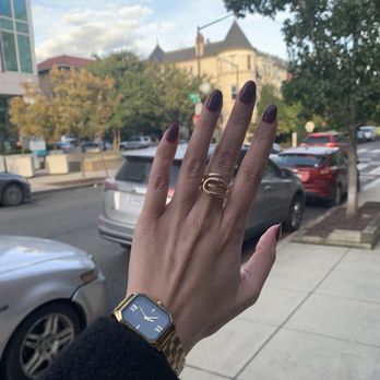 Nails by District - A person's left hand is raised outdoors, displaying dark red nail polish, a gold ring, and a gold watch with a black face. The background features a city street with parked cars, trees, and a historic building with turrets, under a partly cloudy sky. - Nail Salons Near Me - Washington, DC, District of Columbia