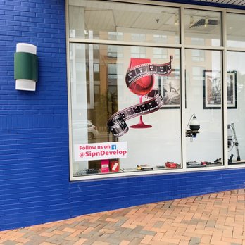 Sip & Develop - The storefront window showcases a large graphic of a wine glass with a film strip wrapped around it, indicating a focus on film or photography. A sign in the window invites viewers to follow the business on Facebook at @SipnDevelop. The interior visible through the glass includes framed pictures on the wall and some equipment on a table, suggesting a creative or media-related business. The exterior wall is painted blue, and the sidewalk is paved with bricks. - Event Photography Near Me - Oxon Hill, Maryland