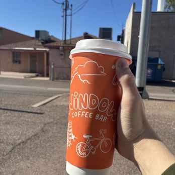 Window Coffee Bar - A person is holding a coffee cup with an orange sleeve from Jindong Coffee Bar, with a white lid, in front of an outdoor urban setting. The background includes buildings, utility poles, and a clear blue sky, indicating a sunny day. The hand is positioned to show the cup prominently, with the sleeve featuring illustrations and text related to the coffee shop. - Coffee & Tea Near Me - Phoenix, Arizona