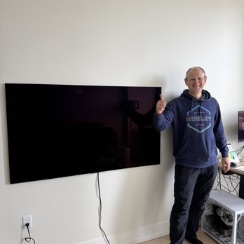 Andrew's TV Installation - A man with a bald head, wearing a navy blue hoodie and dark pants, stands in a room next to a large, wall-mounted flat-screen TV. He is smiling and giving a thumbs-up gesture. The TV is mounted on a plain white wall, with visible cables hanging down. To the right, there is a small desk with electronic equipment and a computer monitor. The room has light-colored wooden flooring and minimal furnishings. - TV Mounting Near Me - Brooklyn, New York
