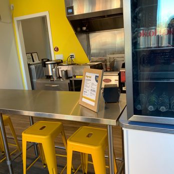 Chee-Bog - Inside - The image shows the interior of a restaurant or cafe with a stainless steel counter and yellow metal stools arranged along it. A beverage fridge containing bottled drinks is visible on the right side. The background includes a yellow wall, a doorway, and a kitchen area with cooking equipment and utensils. The setting appears modern and clean, with a focus on casual dining. - Filipino Near Me - Cohoes, New York