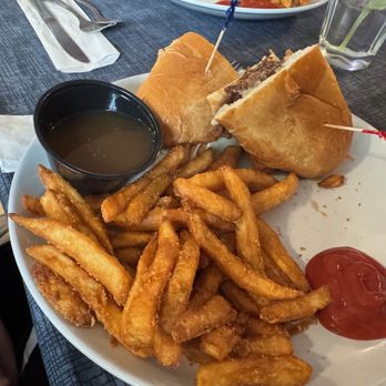 The Brass Lantern - french dip - A plate features a French dip sandwich with beef, French fries, a small cup of gravy, and ketchup, served on a dark table. - Pubs Near Me - Chippewa Falls, Wisconsin