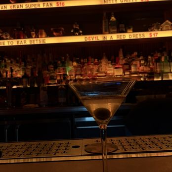 Bar Betsie - martini - A clear martini with an olive is positioned on a bar counter at Bar Betsie in Washington, DC. The background features shelves stocked with various liquor bottles and illuminated signage displaying drink names and prices. The bar has a dark, vintage-inspired decor with warm lighting, and the martini glass is centered in the foreground, highlighting the drink's presentation. - Cocktail Bars Near Me - Washington, DC, District of Columbia