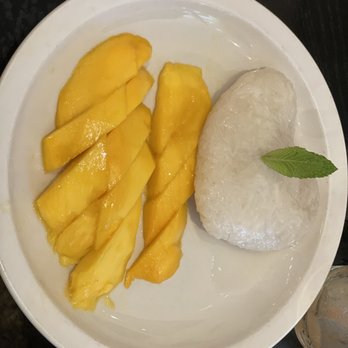 Thai Pavilion - Mango Sticky Rice - The image shows a white plate containing slices of ripe mango and a portion of sticky rice, garnished with a single mint leaf. The mango is cut into several pieces, and the sticky rice is shaped into an oval form. The presentation suggests a traditional Thai dessert, with the mango placed alongside the rice for serving. - Thai Near Me - DeKalb, Illinois