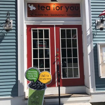 Tea For You - Entrance to Tea for You - The image shows the entrance of "Tea For You," a bubble tea cafe. The storefront has red double doors with white grid-patterned windows, framed by white trim. Above the doors, there is a sign with the business name and offerings such as bubble tea, sandwiches, and salad rolls. A sidewalk sign in front displays contact information and a graphic of a bubble tea cup. The building exterior is painted in blue with decorative lighting fixtures on either side of the entrance. - Bubble Tea Near Me - York, Maine
